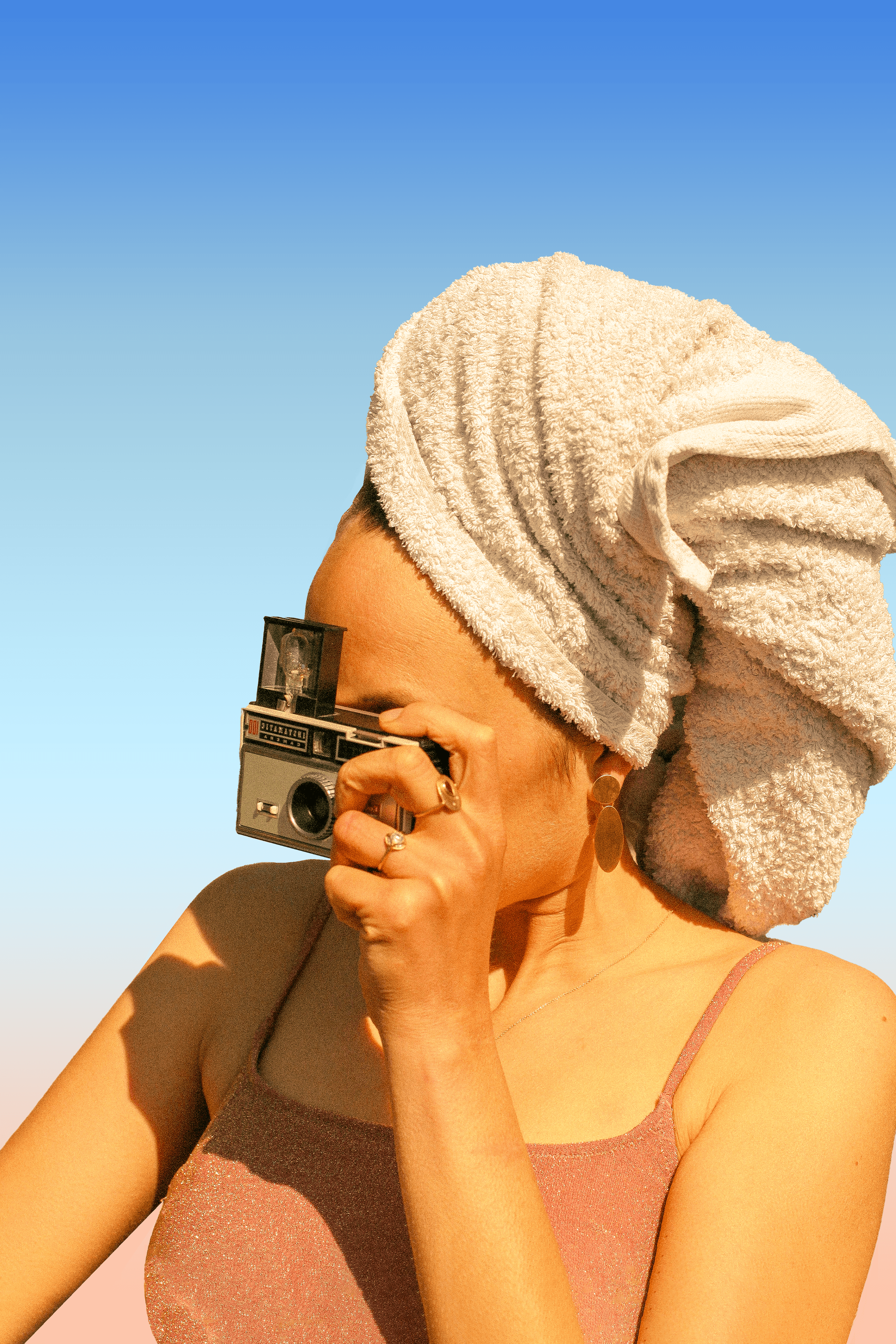 On voit une jeune femme avec une serviette sur la tête, un appareil photo entre les mains, et qui porte des bijoux Louise Vurpas pour Bijouterie Fine - Bijouterie-joaillerie, sur-mesure